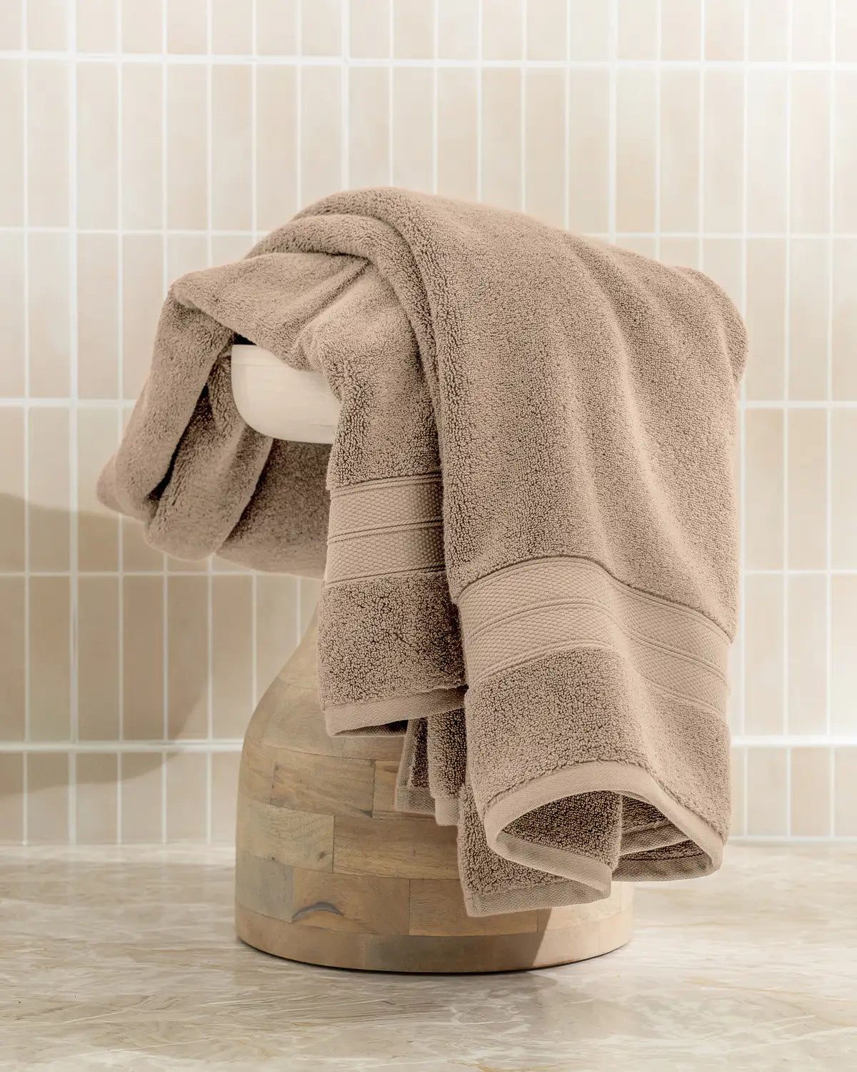 Sandshell modal cotton bath sheet towel draped over a wooden stand against a tiled bathroom wall.