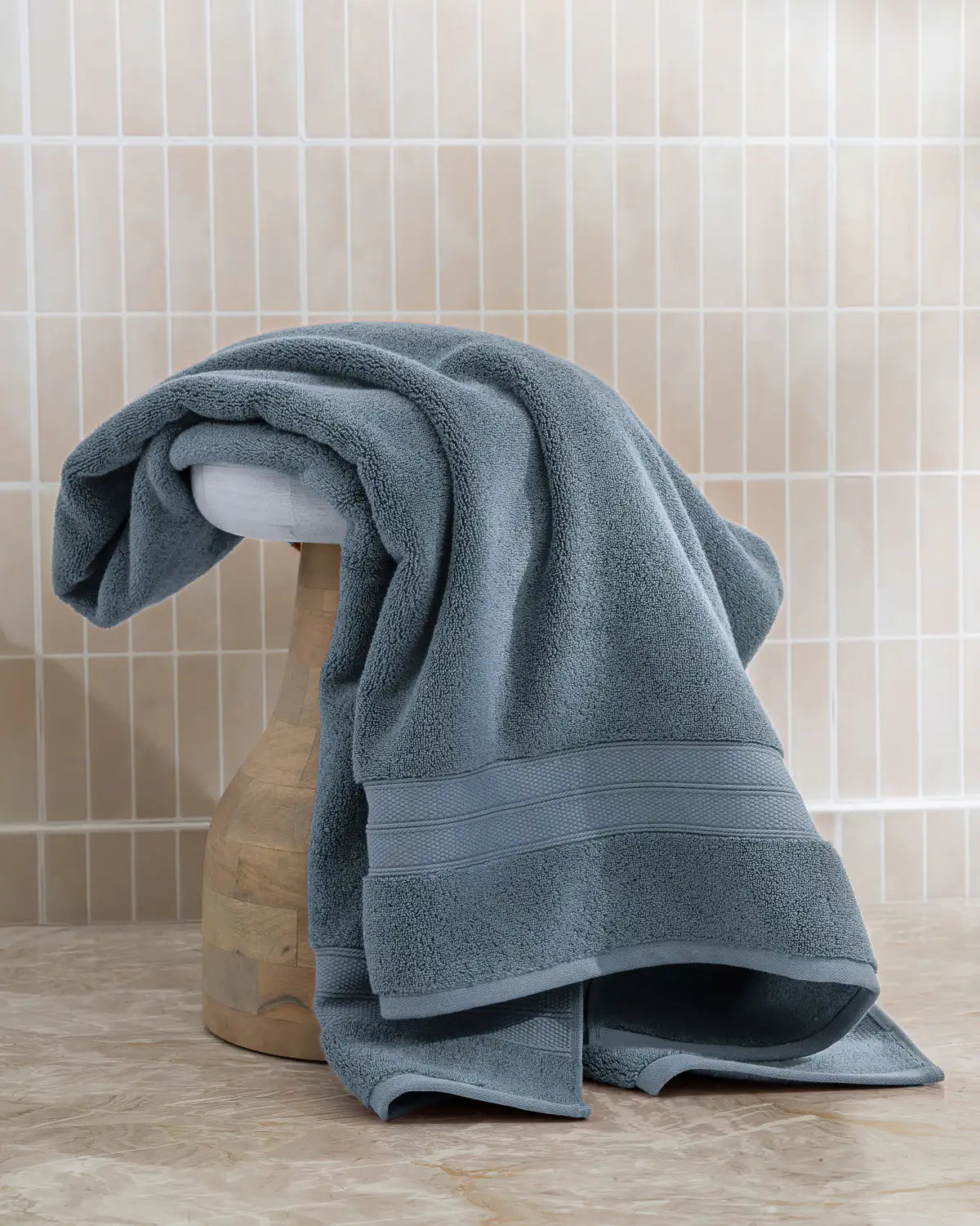 Blue shadow modal cotton towel draped over a wooden stool against a tiled bathroom wall.