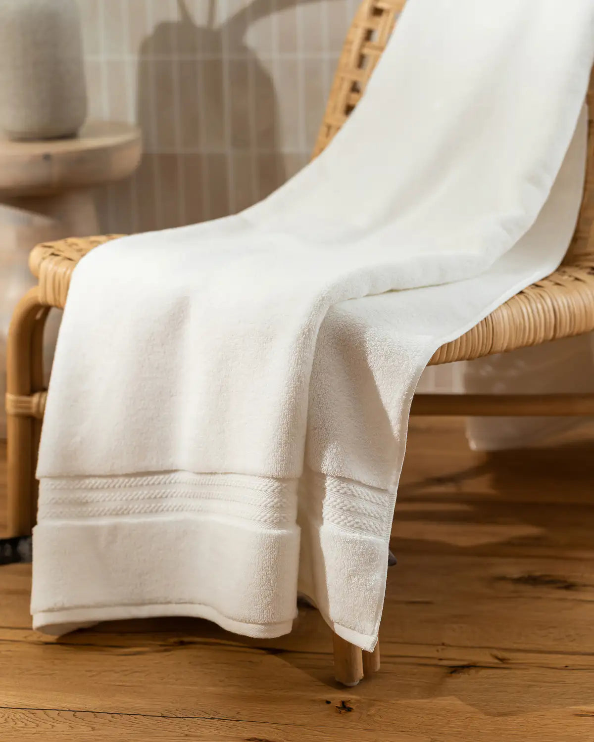 White ultrasoft premium cotton bath towel draped over a wicker chair on a wooden floor.