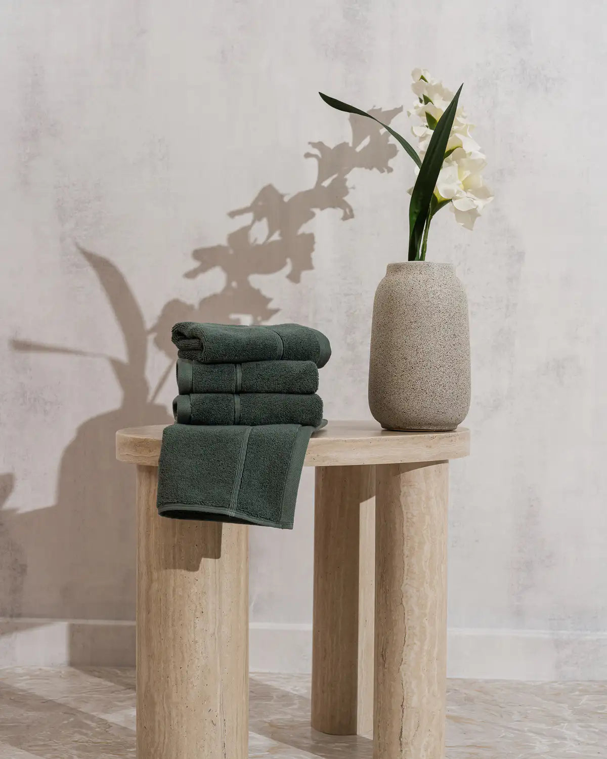 Stack of pine green Turkish bath towels on a wooden stool with a vase containing a white flower against a light gray wall.