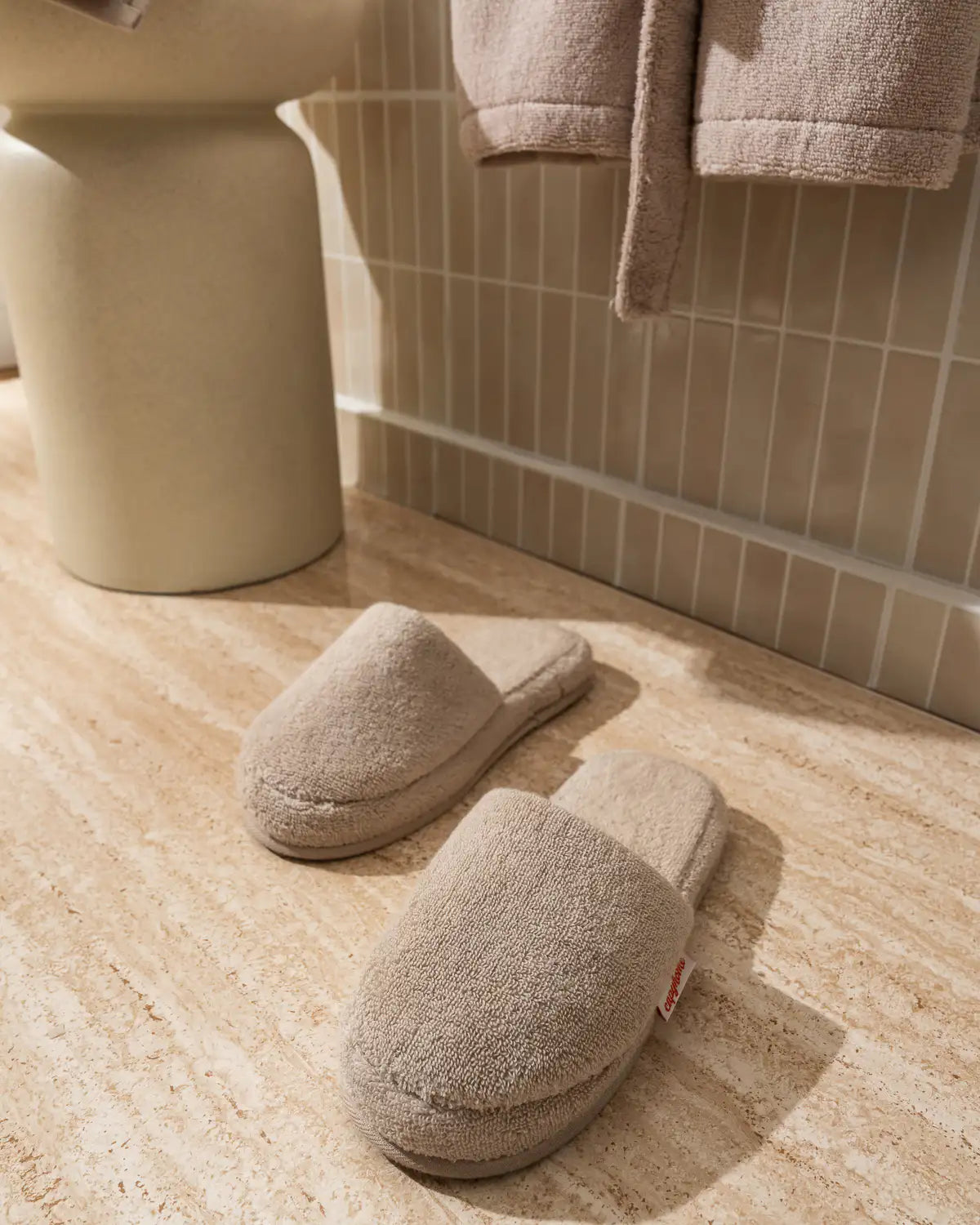 Pair of sandshell UltraSoft Cotton High-Absorbency Slippers on a bathroom floor and with a UltraSoft Premium Cotton High-Volume Bathrobe in the background.