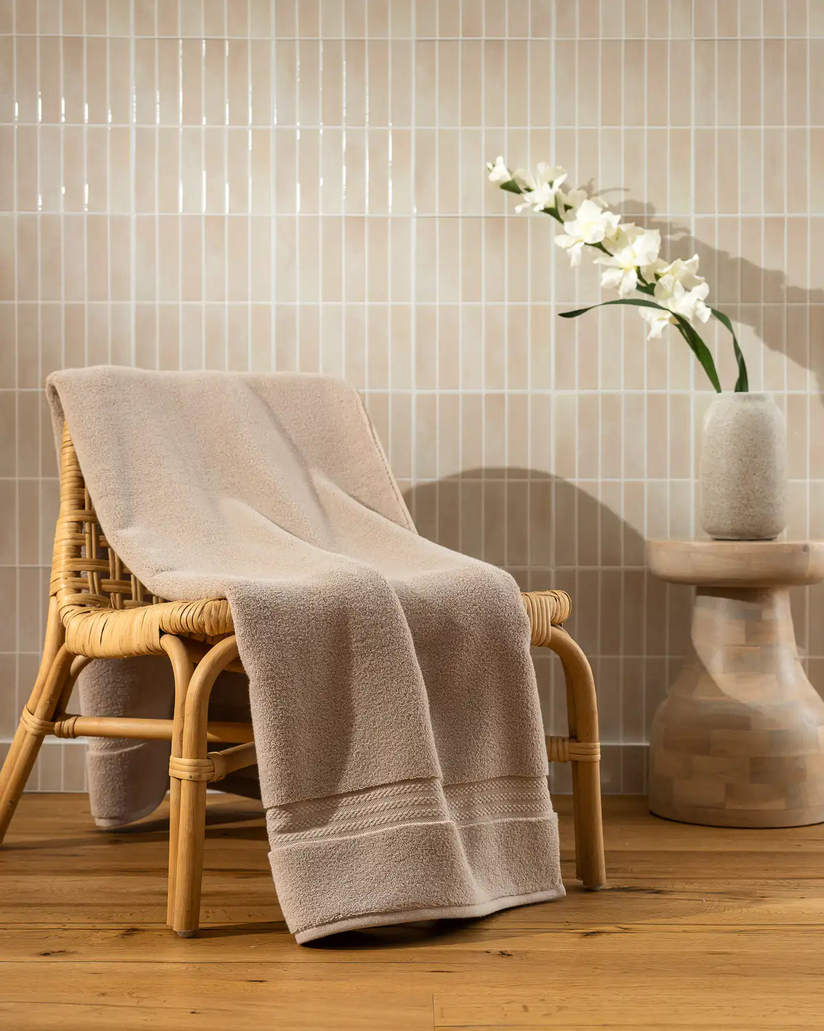 Sandshell ultrasoft premium cotton bath sheet draped over a wooden chair with a vase of flowers on a matching table against a tiled wall.