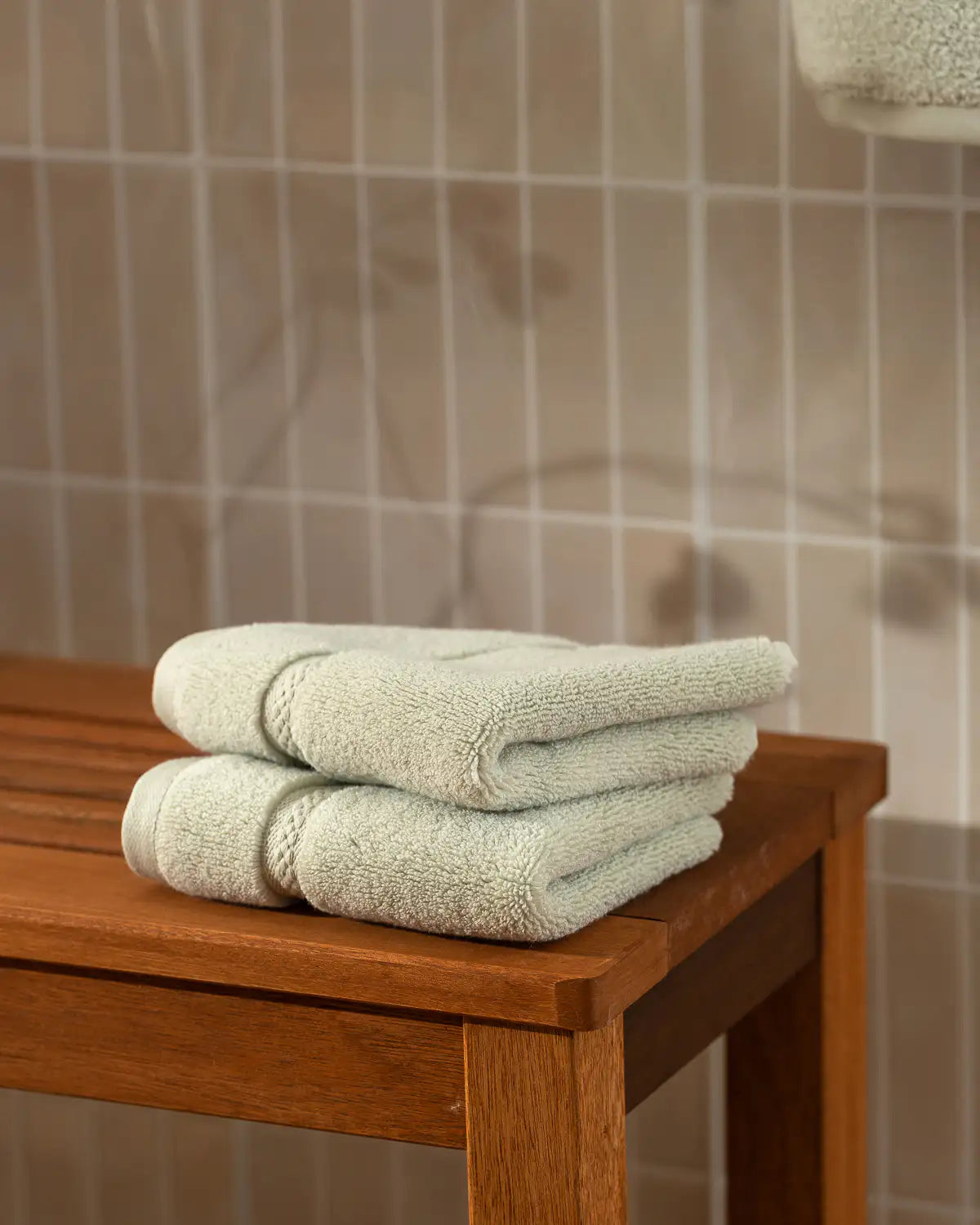 Stack of folded ultrasoft washcloth towels on a wooden bench against a tiled bathroom wall.