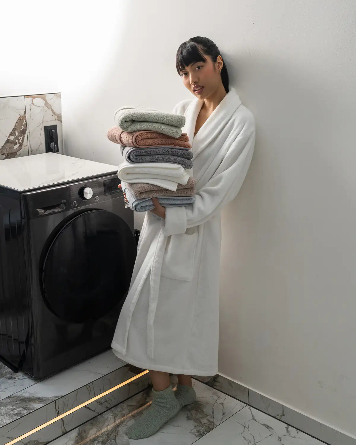 Woman in a white ultrasoft bathrobe holding folded ultrasoft premium cotton towels in a bathroom.